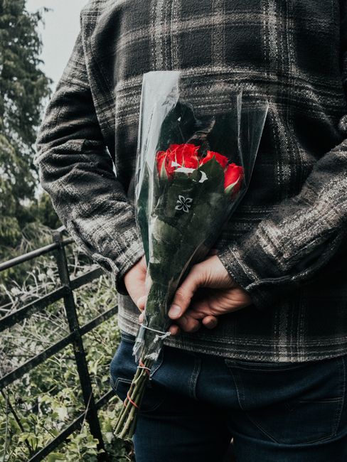 A person wearing a dark plaid jacket holds a bouquet of red and white roses wrapped in clear cellophane, with the stems neatly gathered and secured with a small ribbon. The bouquet features fresh, vib