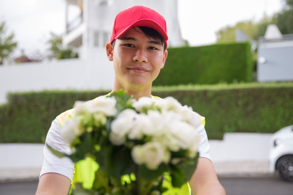 A male flower delivery courier wearing a red cap and a high-visibility vest holds out a bouquet of white flowers towards the camera. The bouquet features fresh, tightly packed petals and green stems, 
