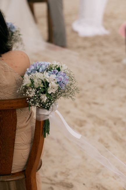 A bridal bouquet held by a bride seated on a wooden chair at a beach wedding, featuring a delicate arrangement of white and light purple flowers, including roses and baby's breath, wrapped with a whit