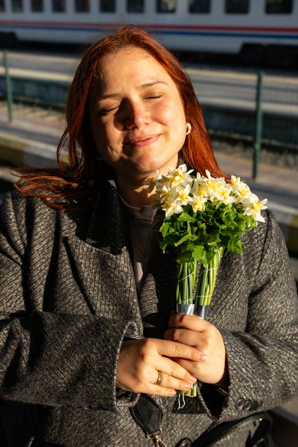 A woman with closed eyes and a gentle smile is holding a clear glass vase filled with fresh white and yellow daffodils, their petals appearing soft and vibrant, with bright green stems and leaves. She