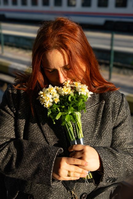 A woman with vibrant red hair, dressed in a dark grey textured coat, holds a bouquet of fresh white and yellow daffodils. The bouquet is arranged with green stems gathered together and wrapped in a cl