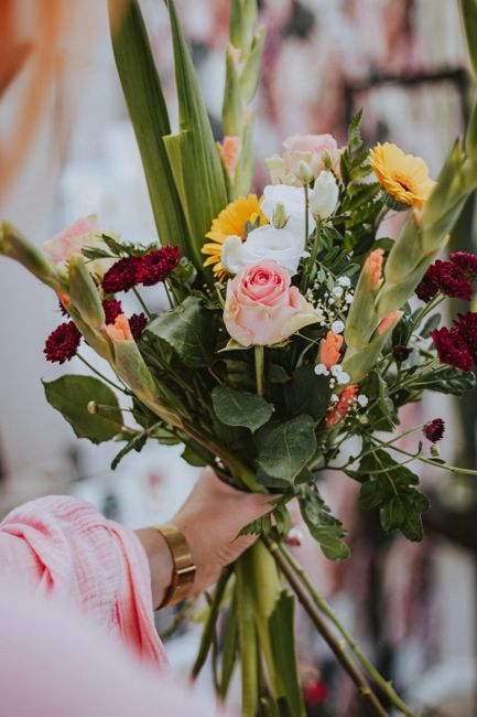 A hand holding a vibrant bouquet of fresh flowers. The arrangement includes light pink roses, white calla lilies, yellow daisies, and deep red chrysanthemums, complemented by greenery and long, green 