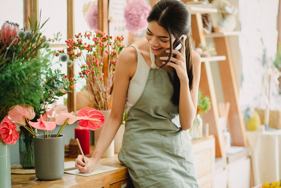 A young woman with long dark hair, smiling, and wearing a light-colored sleeveless top and a beige apron, is standing in a well-lit florist shop. She is holding a telephone receiver with her right han