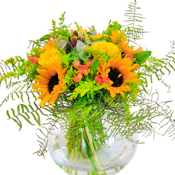 The immediate impression of this lavish bouquet is one of pure sunlit delight, carefully crafted to reflect the seasonal vibrance of Sands End. Vivid sunflowers, bold and inviting, radiate with glowing yellow-orange petals that seem to pour warmth into any room. Beside these beacons of cheer, velvety yellow garden roses unfurl tier by tier, each petal folding into the next with the promise of softness and classic beauty. Lush green foliage spills outward, anchored by tumbling ferns and slender sprigs of goldenrod that carry clusters of tiny yellow flowers, evoking the untamed abundance of local riverside gardens in peak bloom. Glossy Hypericum berries in brilliant red are thoughtfully tucked throughout, their pop of colour giving the bouquet a modern, energetic twist. Wisps of eucalyptus bring a calming aroma, and their frosted greenery elegantly contrasts the sun-warmed palette. The round, crystal-clear vase on which the bouquet rests reveals the interlacing stems bathed in fresh water, adding architectural grace and clarity to the arrangement. Perfect as an uplifting centrepiece for family celebrations, milestone birthdays, or anniversaries in and around Sands End, this composition-lovingly arranged by Flower Delivery Sands End-embodies joy, tradition, and the fresh spirit of West London floristry.